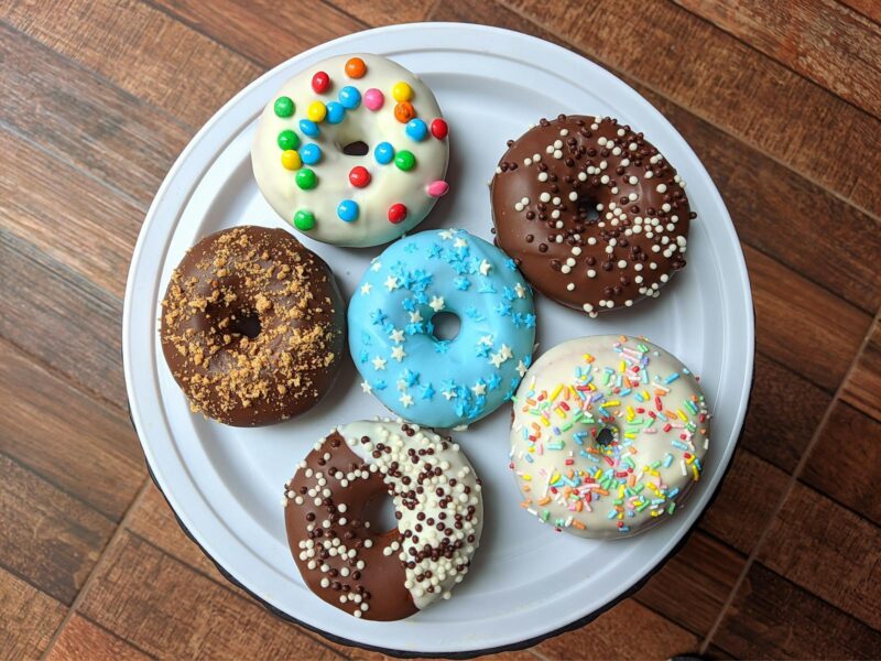 six multi-colored donuts on a white plate sitting on a brown tabletop