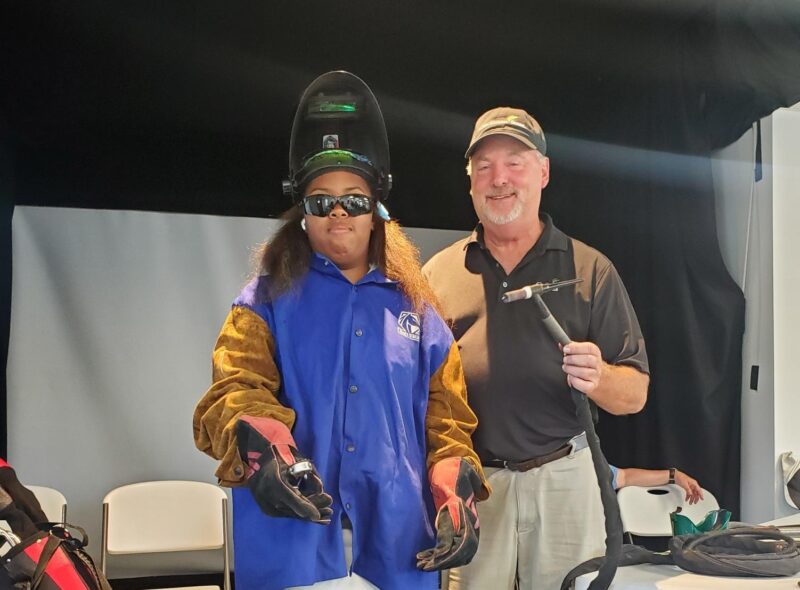 Welding instructor Bruce Scherer with Reaching Out For Kids camp participant dressed in welding helmet and gloves